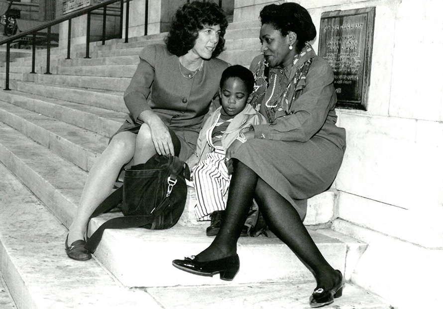 Two women sit on stone steps, engaged in conversation, while a young child sits between them looking down. All three wear formal attire. A bag rests beside one woman, and a plaque is visible on the wall behind them.