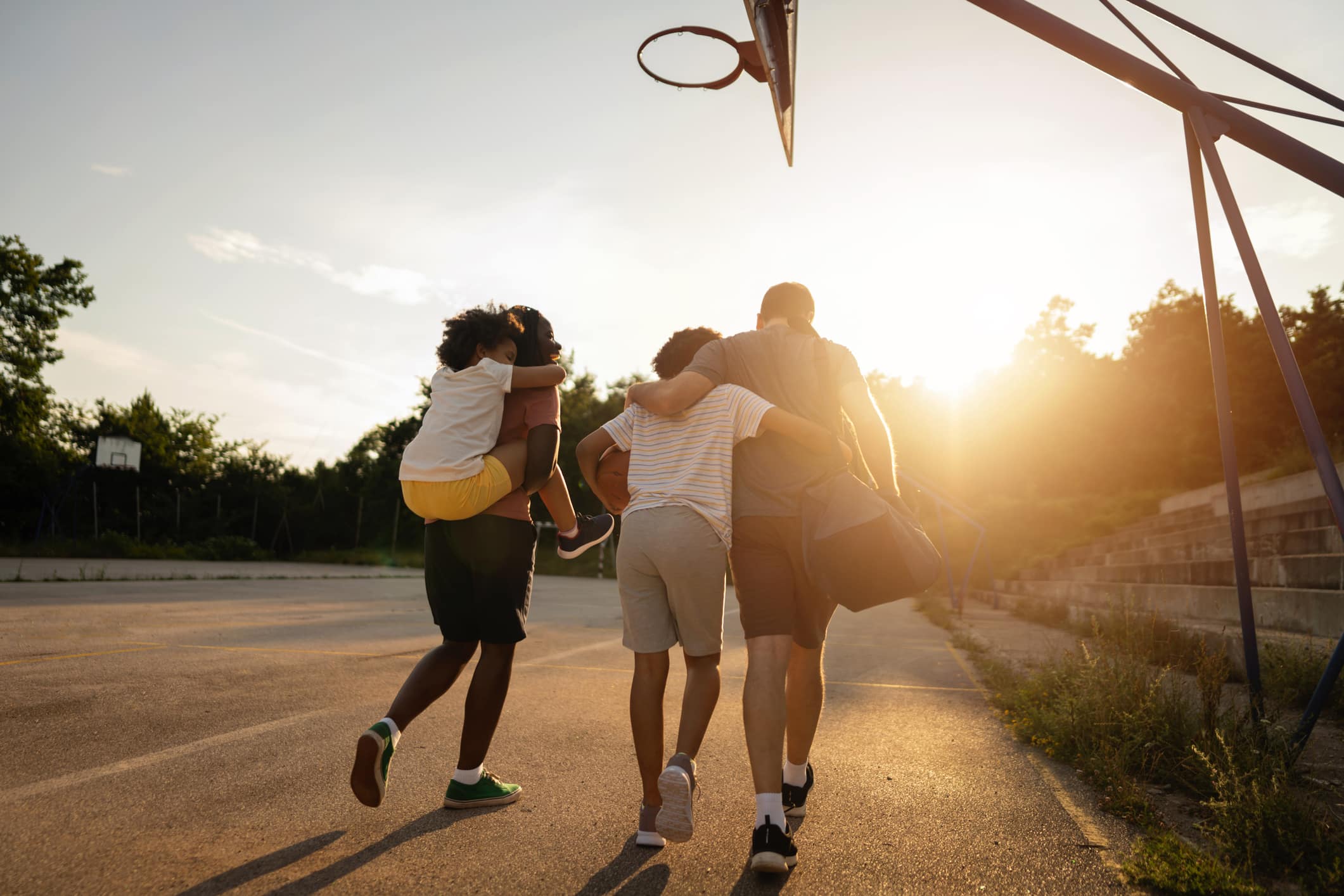 Three people walk on an outdoor basketball court at sunset, with one carrying another on their back and the third holding a sports bag. The court is empty, with trees and bleachers visible in the background.