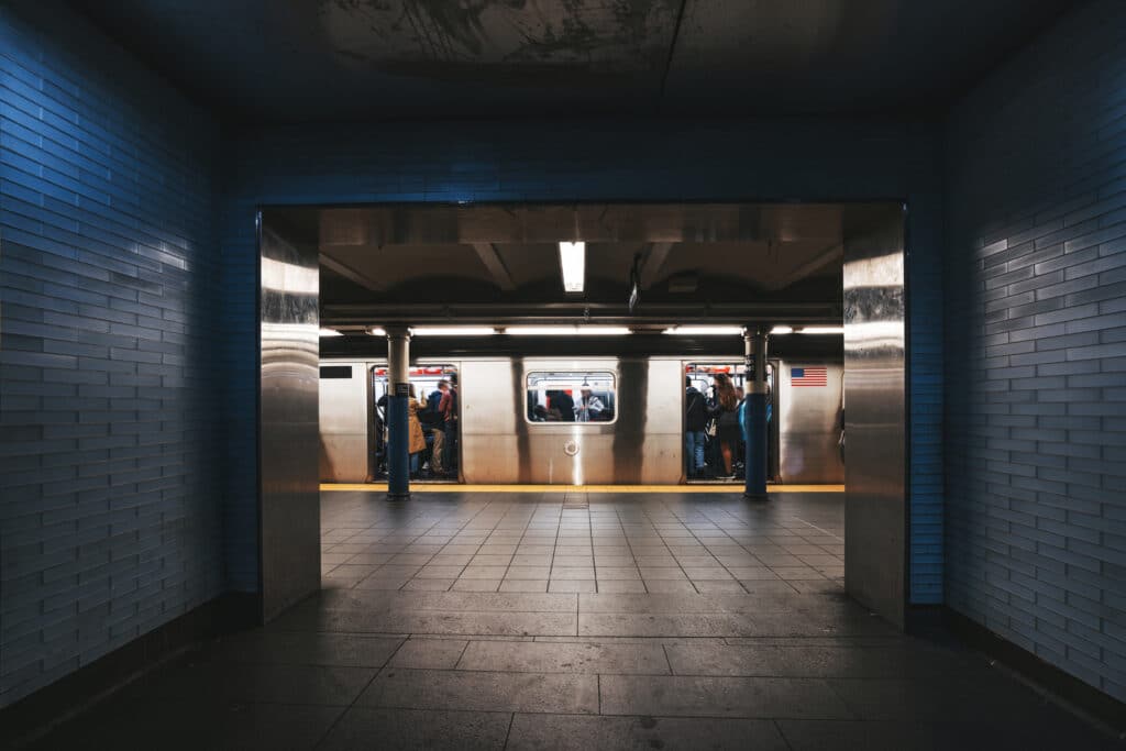A subway platform seen through a tiled hallway, with people standing and sitting inside a stopped train. The trains doors are open, and some passengers are entering or exiting. The lighting is dim and the scene appears urban.