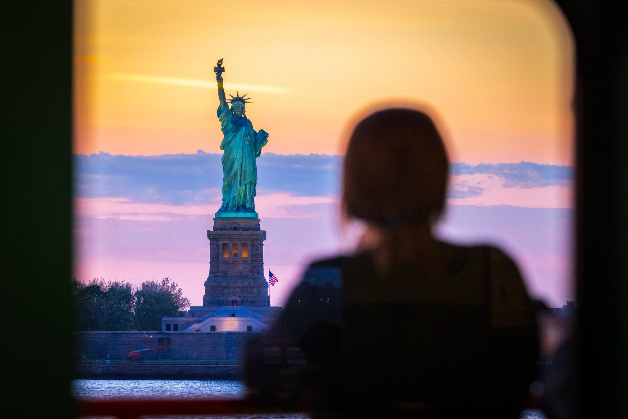 A person is silhouetted in the foreground while looking at the Statue of Liberty at sunset, with the sky displaying hues of orange, pink, and purple. The statue stands on its pedestal with a flag in the background.