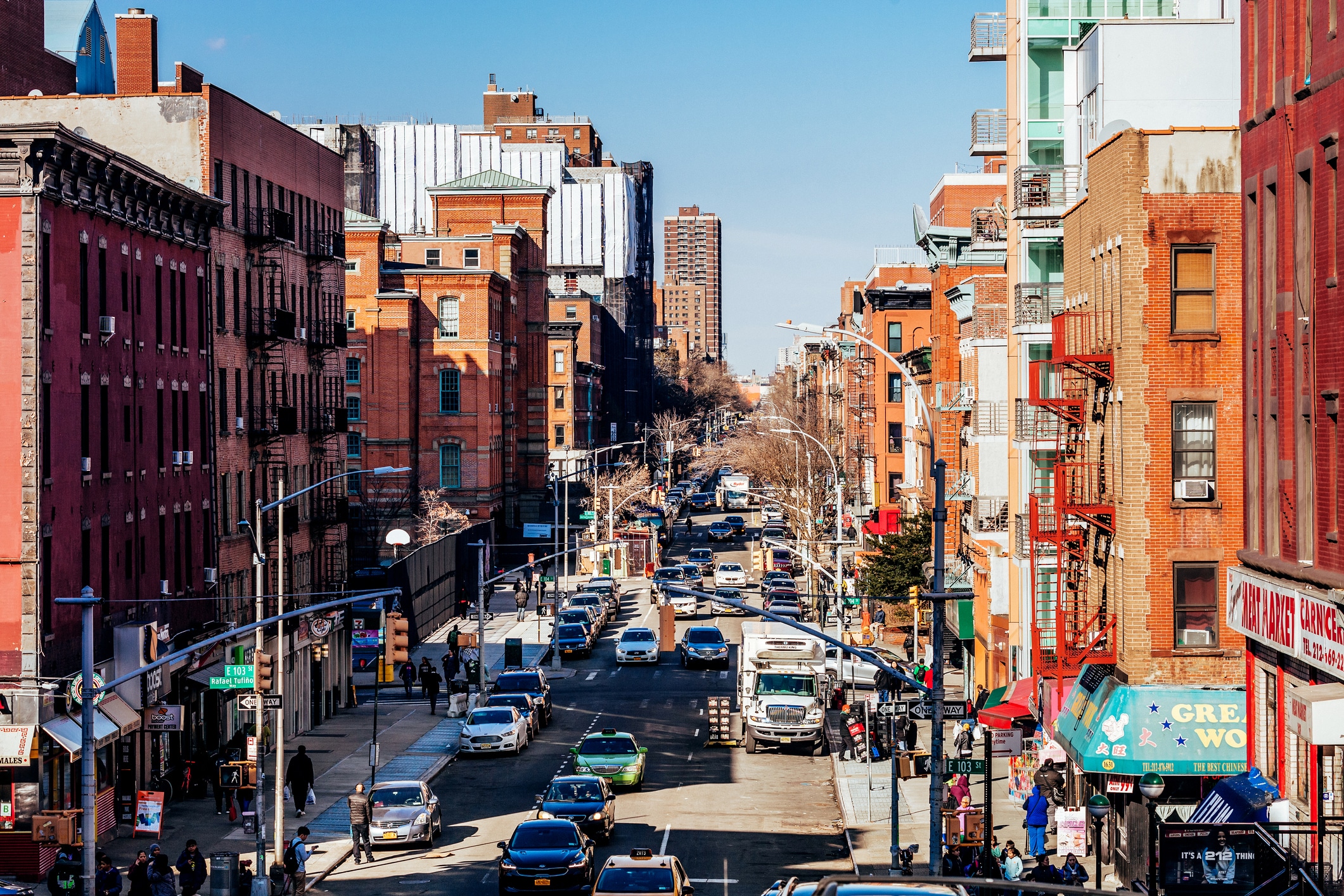 A busy city street lined with red brick buildings, cars, and pedestrians. The street stretches into the distance with traffic and shops visible on both sides under a clear blue sky.