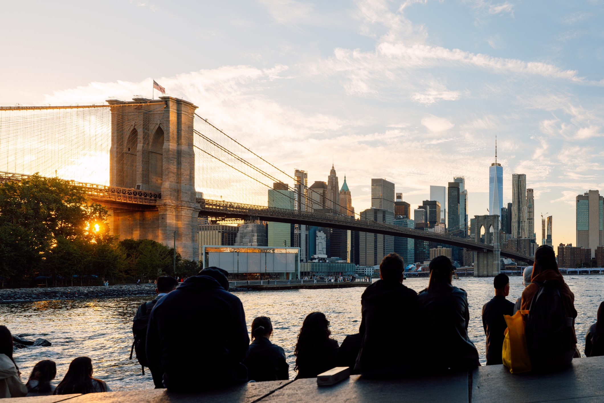 People sit by the water at sunset, facing the Brooklyn Bridge and the Manhattan skyline. The sun is partially hidden behind the bridge, casting a warm glow over the city buildings and clouds.