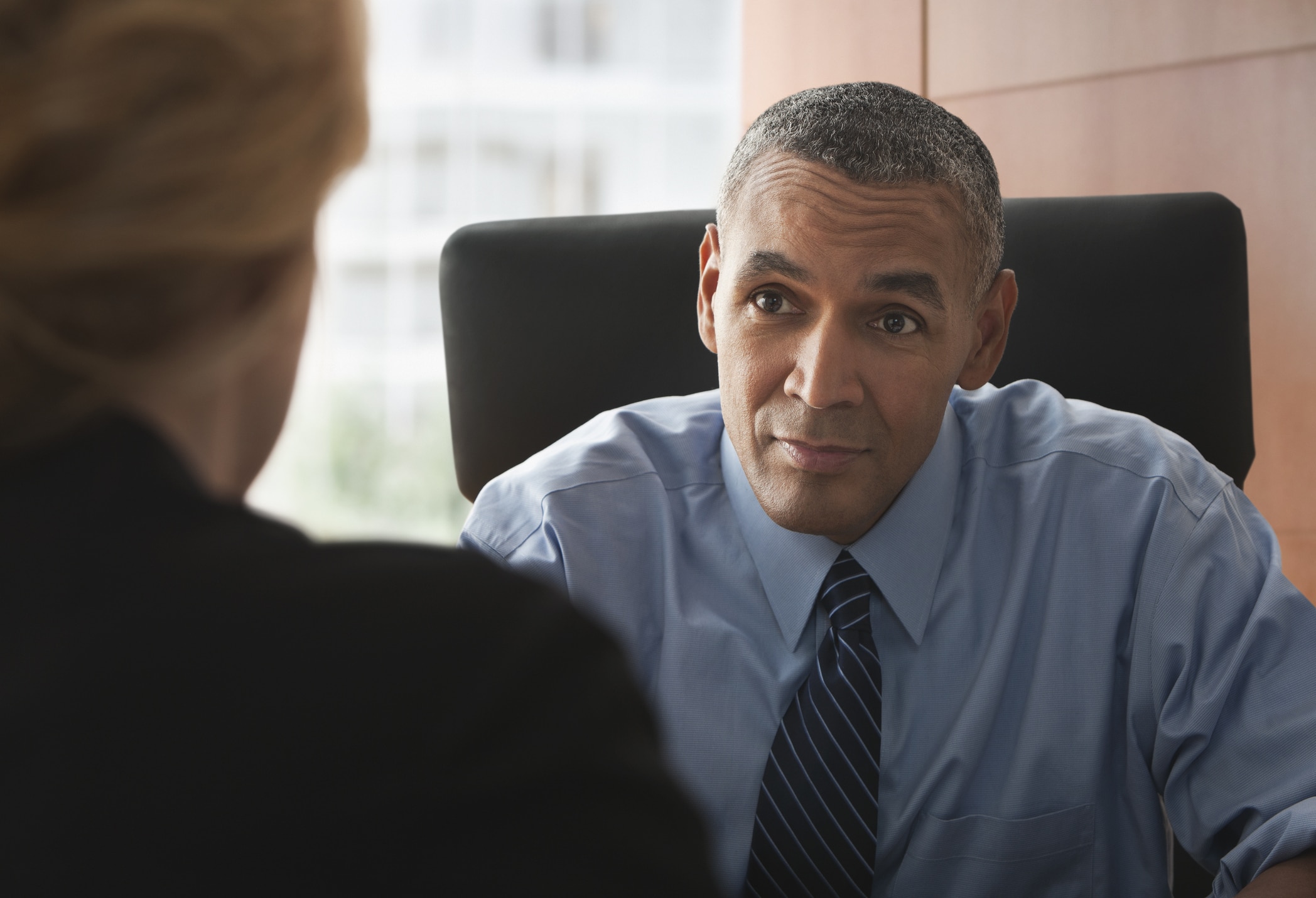 A man in a blue shirt and striped tie sits at a desk, attentively listening to a woman whose back is to the camera. Office setting with a window in the background.