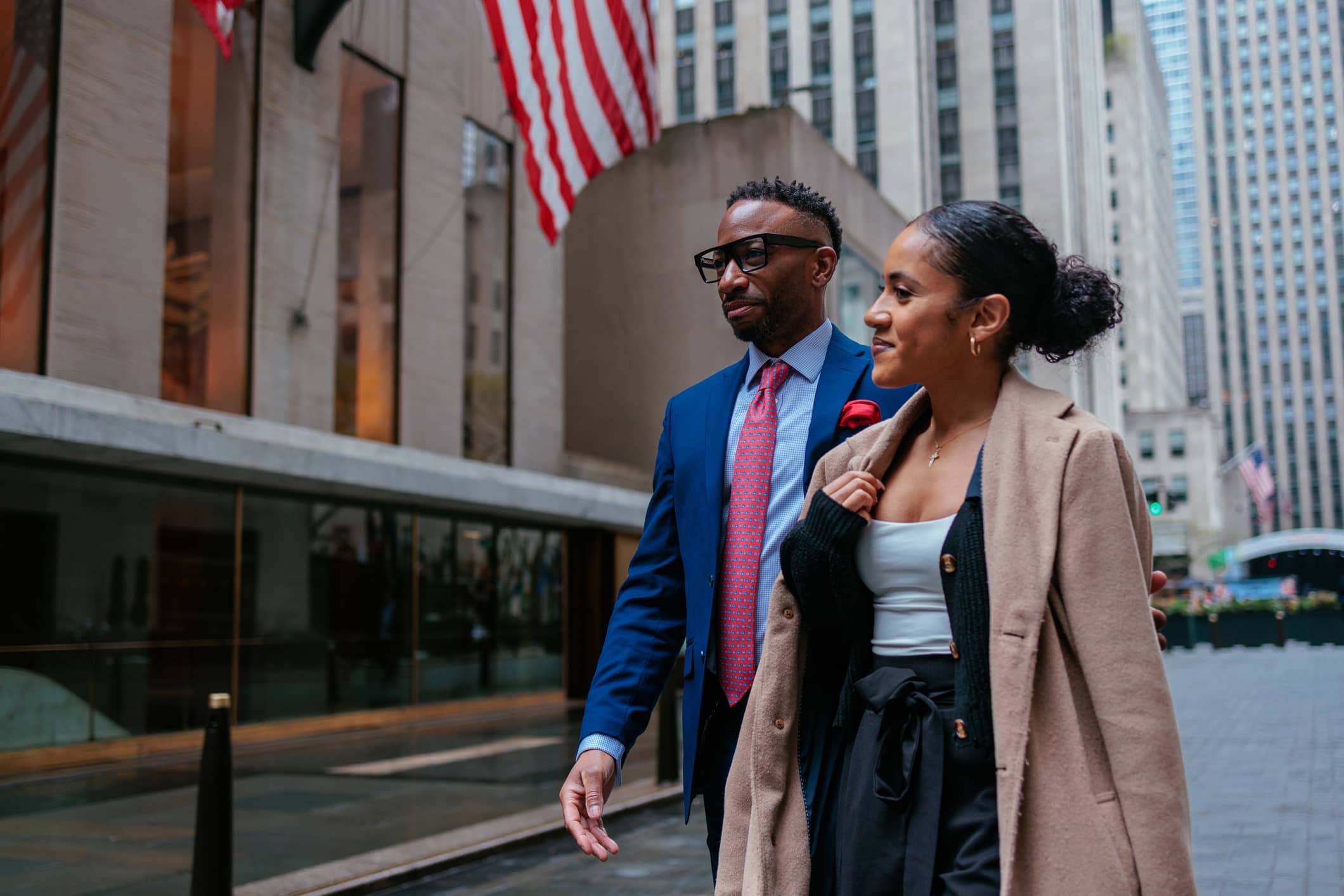 Two people walk side by side on a city sidewalk near tall buildings, both dressed in business attire. American flags hang on the building in the background. The scene appears to be in an urban downtown area.