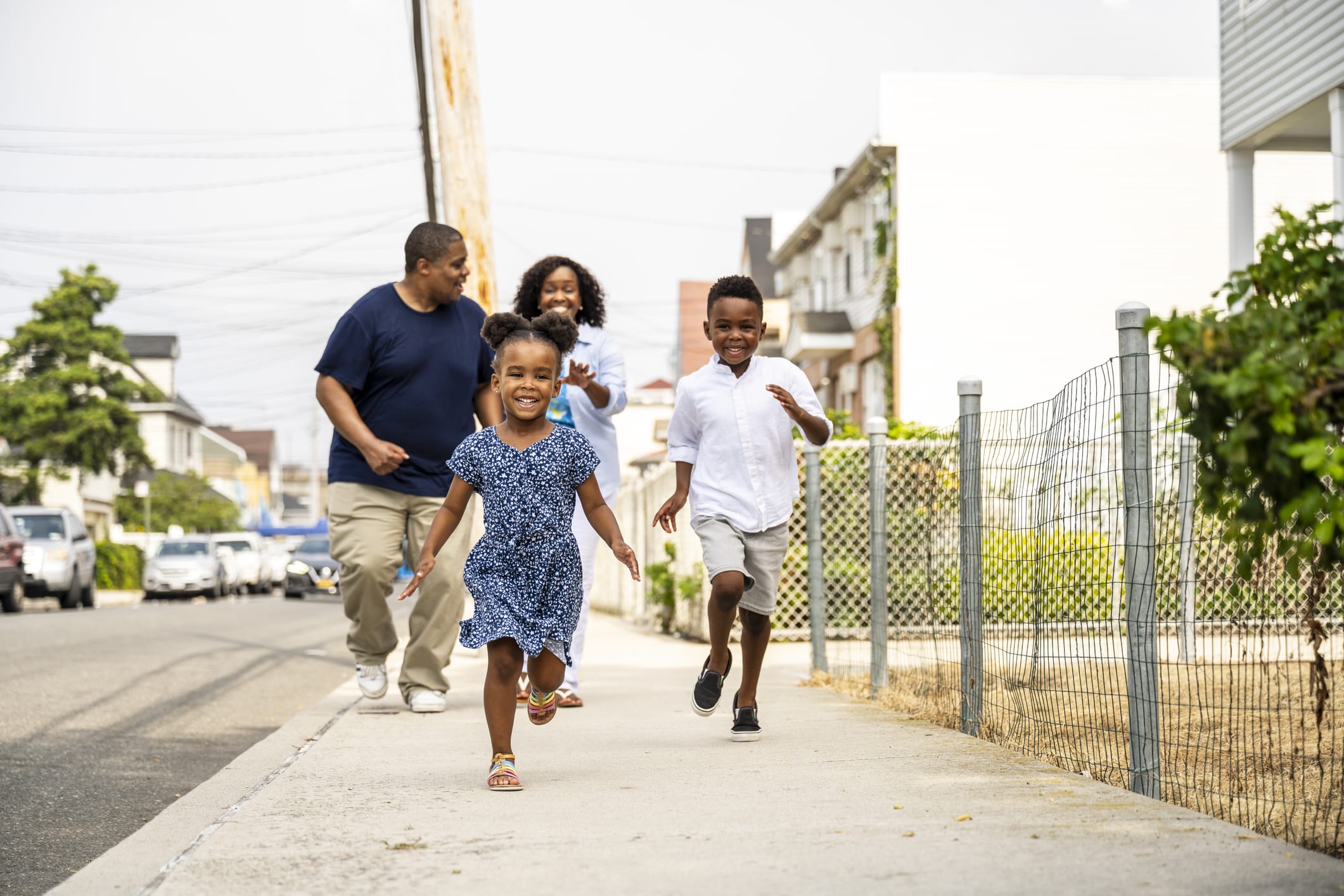 Two children run on a sidewalk while two adults walk behind them. The group is outdoors in a residential area with houses, fences, and parked cars visible along the street.