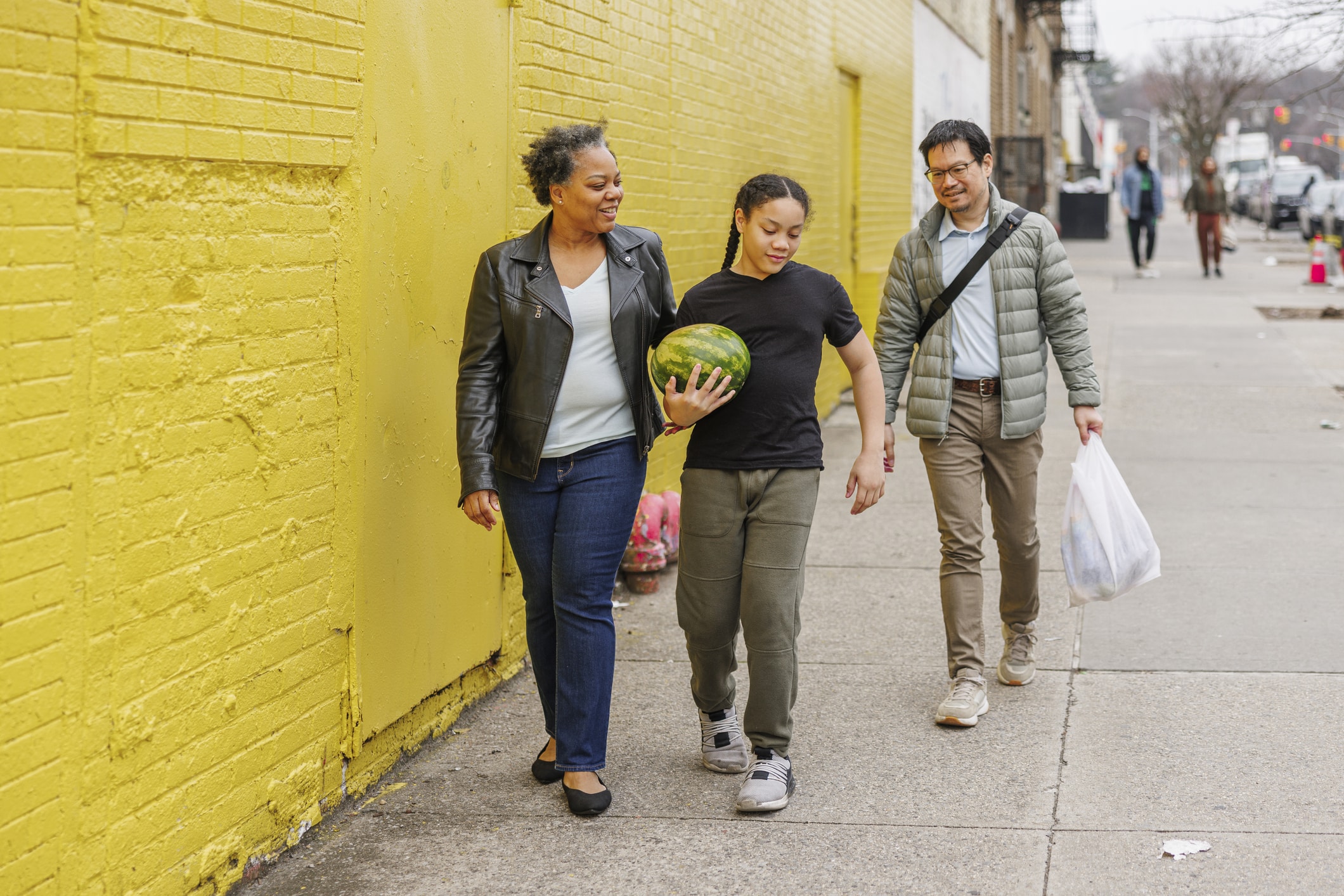 Three people walk on a city sidewalk next to a yellow wall. A woman and a girl, who is carrying a watermelon, walk ahead and smile. A man follows behind carrying two plastic bags.