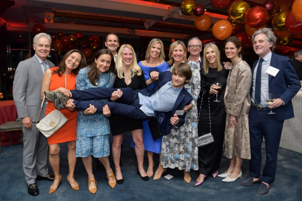 A group of people pose indoors at an event. Most are smiling at the camera; two women lift a boy in a suit. Colorful balloons and red lighting decorate the background. Some hold drinks, and everyone wears formal or semi-formal attire.