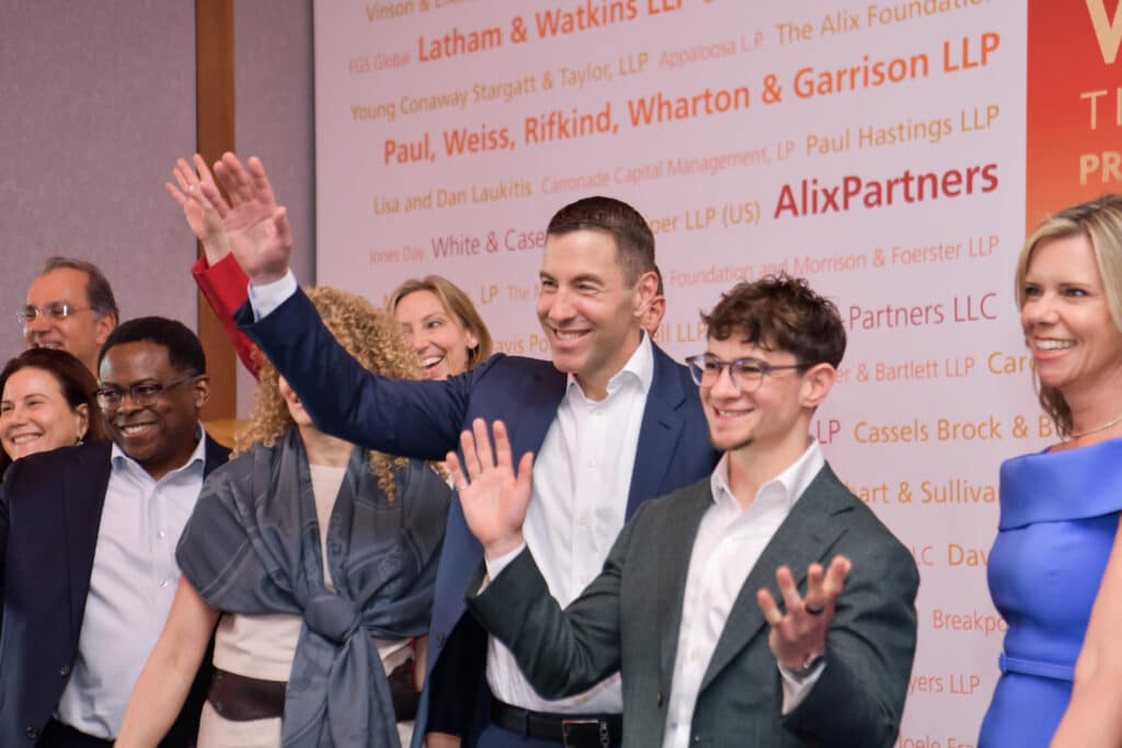 A group of people in business attire stands smiling and waving in front of a wall with multiple law firm names and logos printed on it.