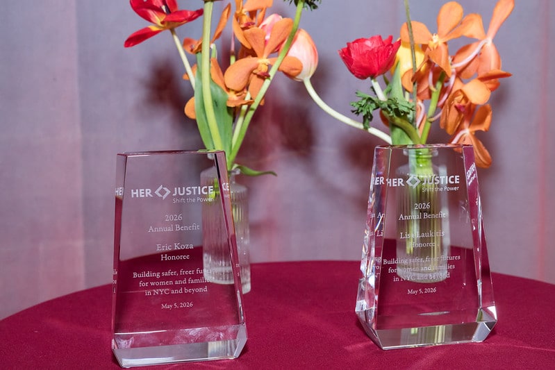 Two crystal awards on a round red tablecloth, with flower arrangements in the background. The awards are engraved with text from the Her Justice 2016 Annual Benefit honoring two recipients.