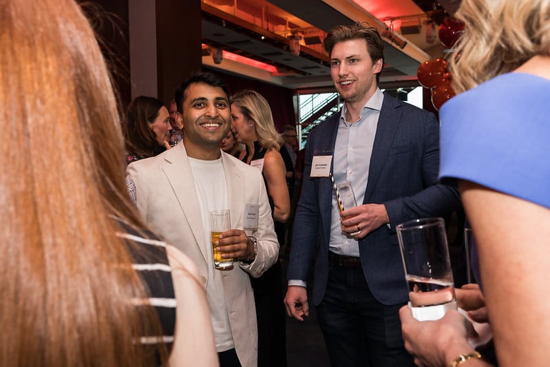 Two people in business attire hold drinks and talk at an indoor event. Other attendees are visible in the background. The setting has warm lighting and some balloons in the back.