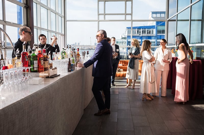A group of people in business and formal attire gather by a bar with bottles and glasses inside a bright room with large windows and views of buildings outside.