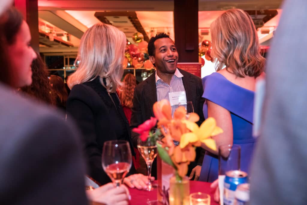 Four people stand around a table with drinks and flowers, talking at an indoor event with warm lighting. One man in a suit is smiling, while the others are engaged in conversation.