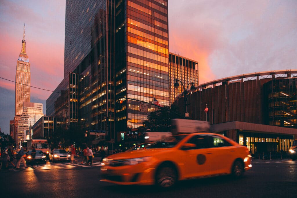 A yellow taxi drives through a busy city intersection at sunset, with tall glass buildings and the Empire State Building visible in the background under a pinkish sky.