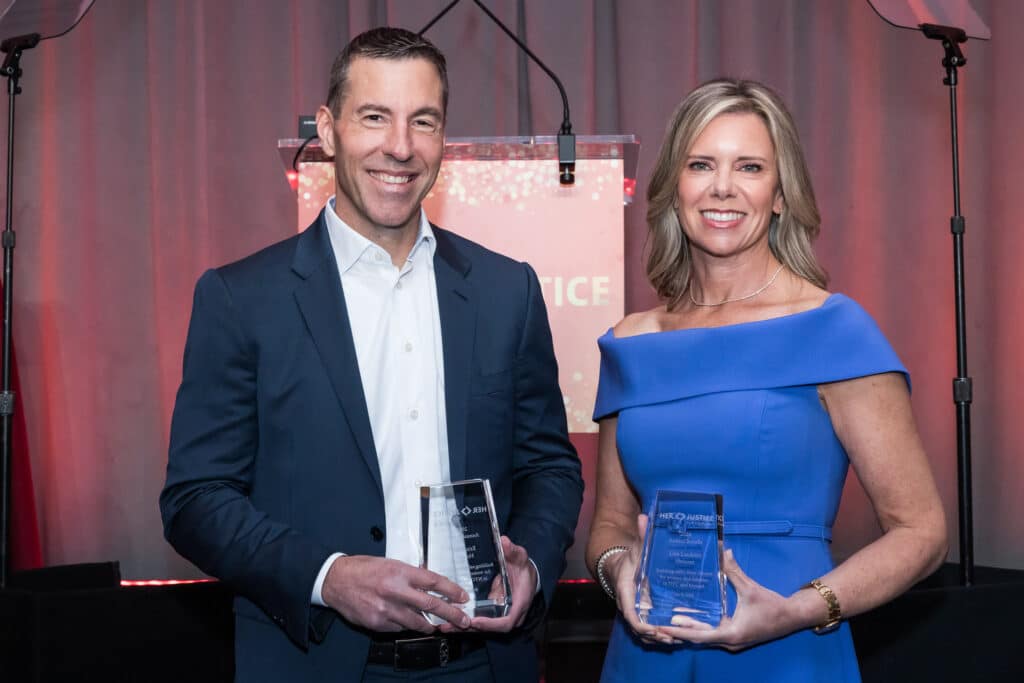 A man in a dark suit and a woman in a blue dress stand side by side, smiling and holding glass awards. A red, lit-up backdrop and microphone are visible behind them.