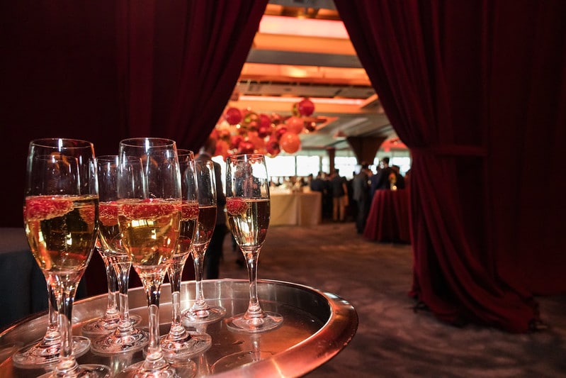 A tray with five champagne glasses garnished with berries is in the foreground. Deep red curtains frame the scene, and a reception with guests and red balloon decorations is visible in the background.