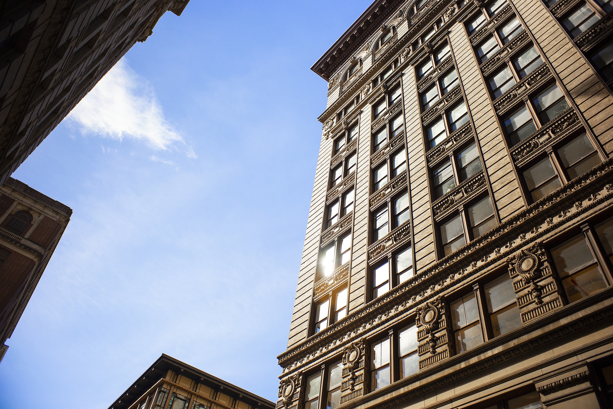 A tall, ornate building with large windows and decorative architectural details is shown from a low angle against a blue sky, with portions of other buildings visible on either side.