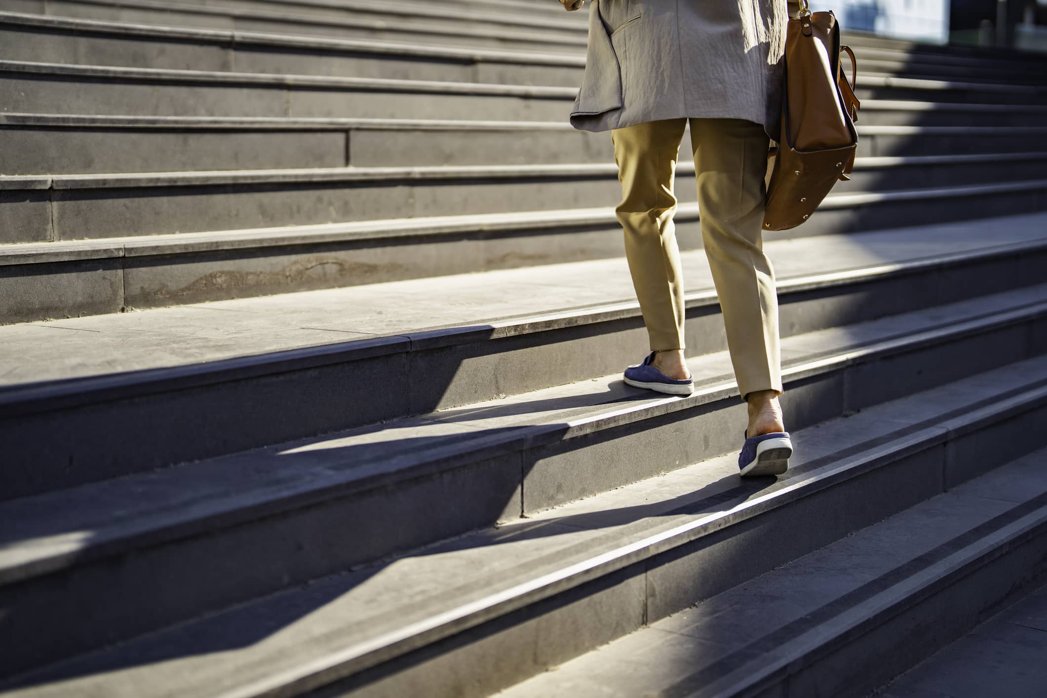 A person wearing tan pants and loafers walks up concrete steps outdoors, carrying a brown leather bag. Only their lower body is visible in the sunlight.