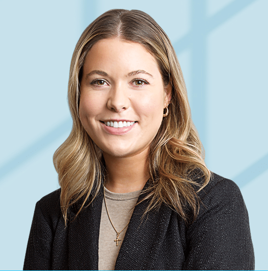 A woman with shoulder-length blonde hair, wearing a black blazer, beige top, and a cross necklace, smiles in front of a light blue background with soft shadows.
