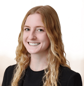 A young woman with long, wavy blonde hair is smiling. She is wearing a black blazer over a light top. The background is plain and bright.