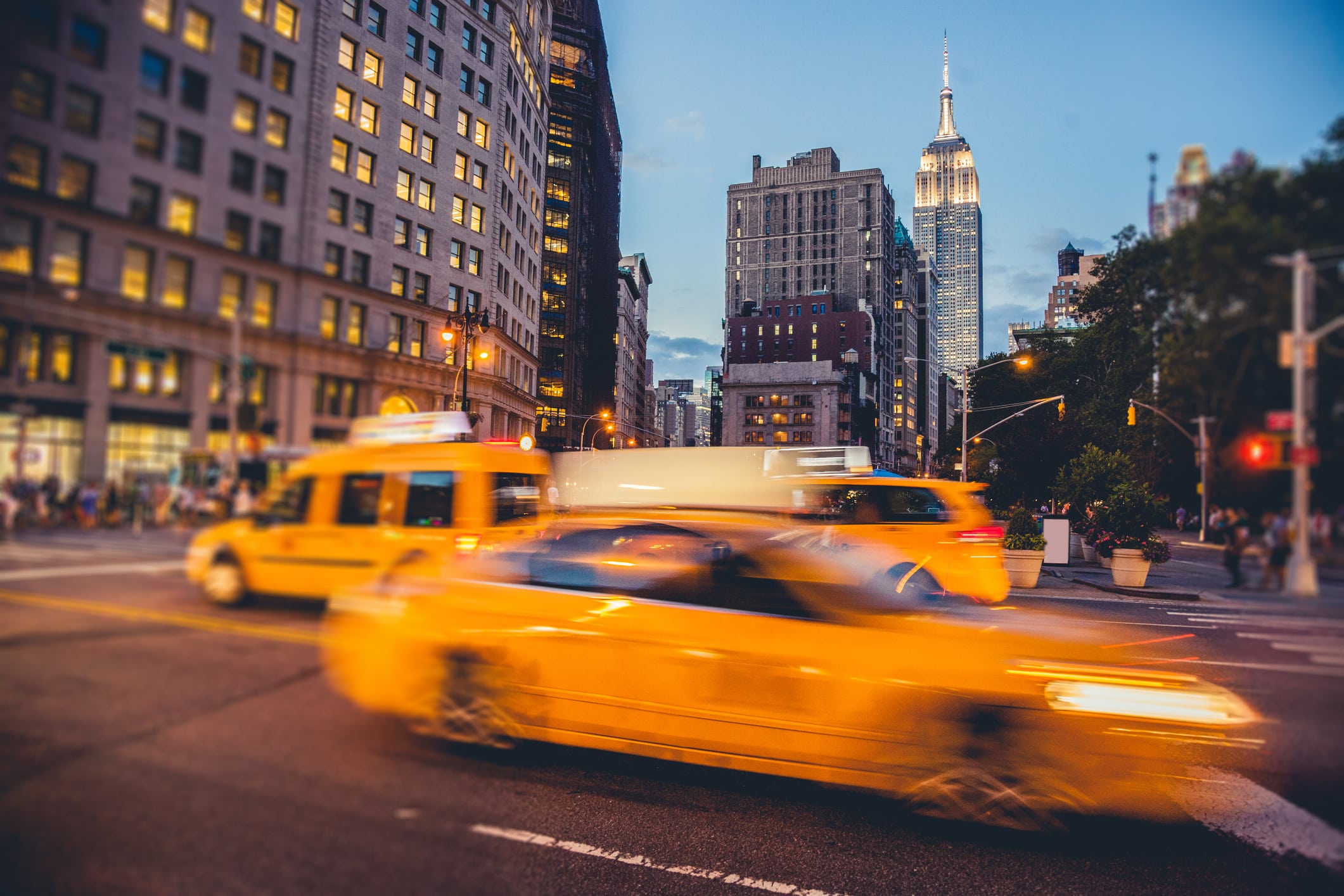 Yellow taxis drive through a busy city intersection at dusk with tall buildings, including the Empire State Building, in the background. The streetlights are on and the sky is starting to darken.