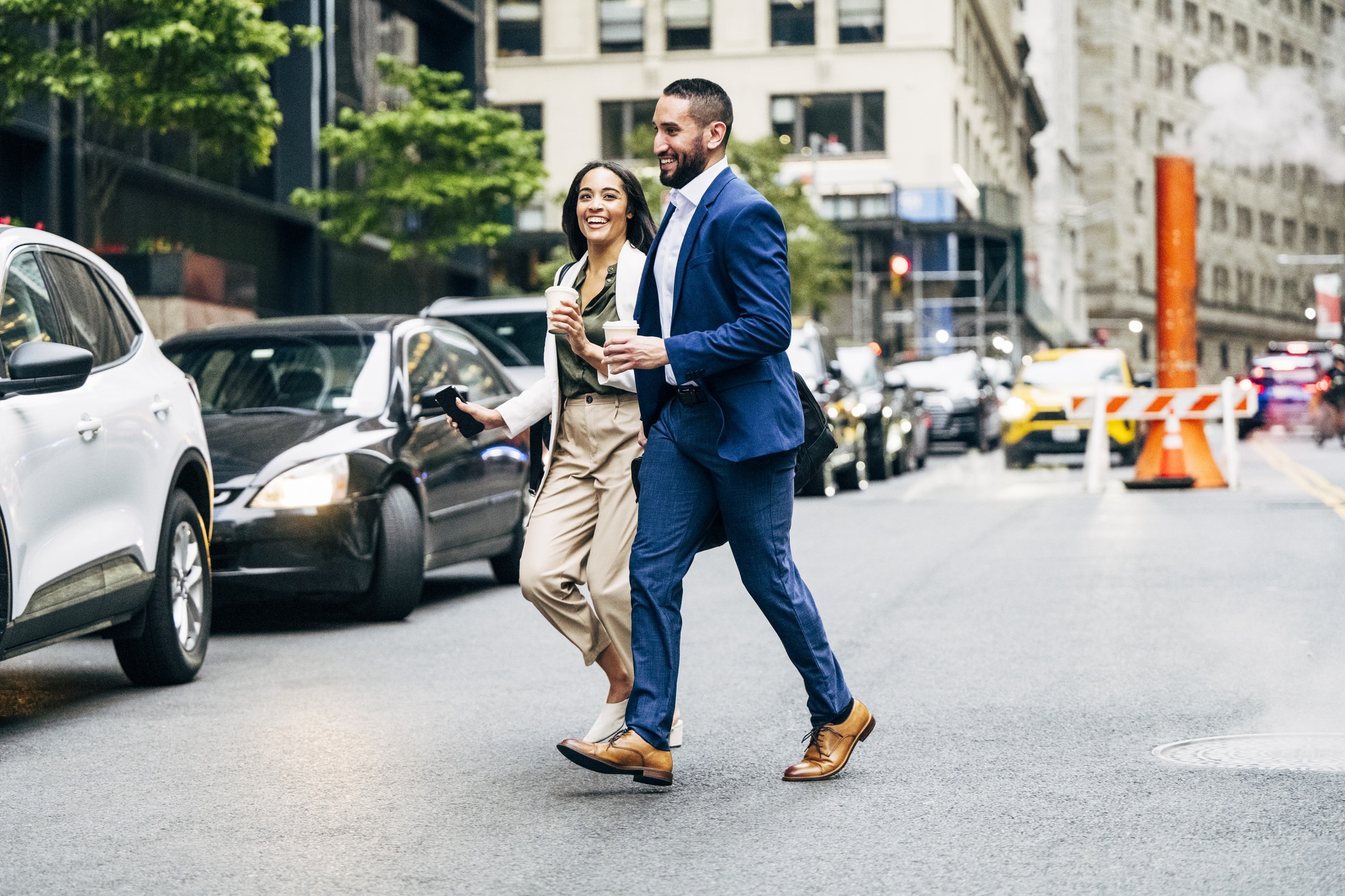 Two people in business attire cross a city street, holding coffee cups and smiling. Cars are stopped nearby, and tall buildings and street construction barriers are visible in the background.