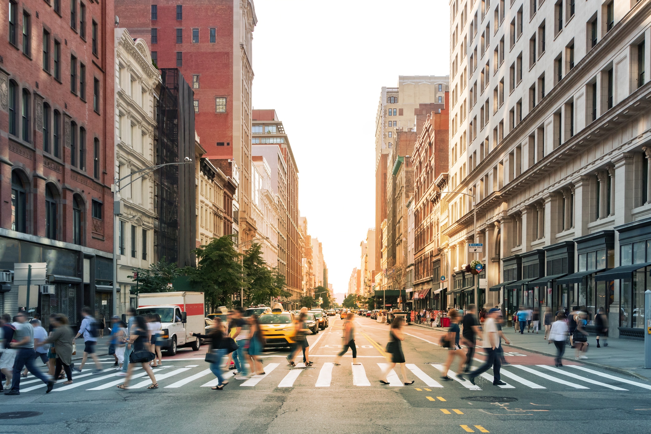 A busy city intersection with many people crossing the street, blurred in motion. Tall buildings line both sides of the street, and cars, including a yellow taxi, wait at the traffic lights.