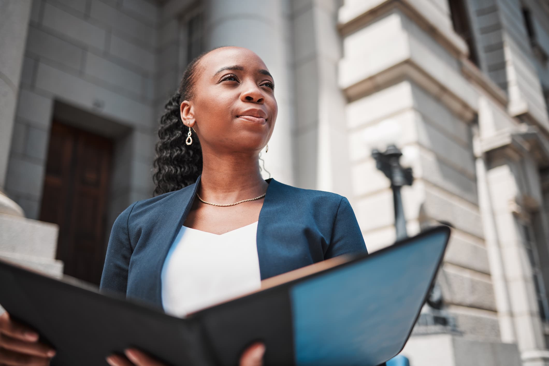 A woman in business attire stands outside a stone building, holding an open folder and looking ahead. The background shows architectural details and steps typical of a government or courthouse building.
