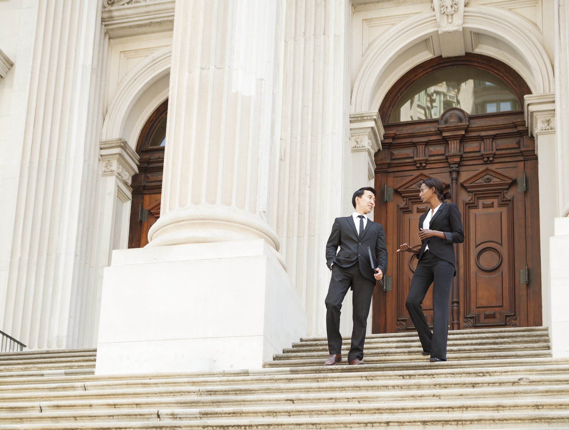 Two people in business attire walk down the steps of a large white building with columns and wooden doors, engaged in conversation.