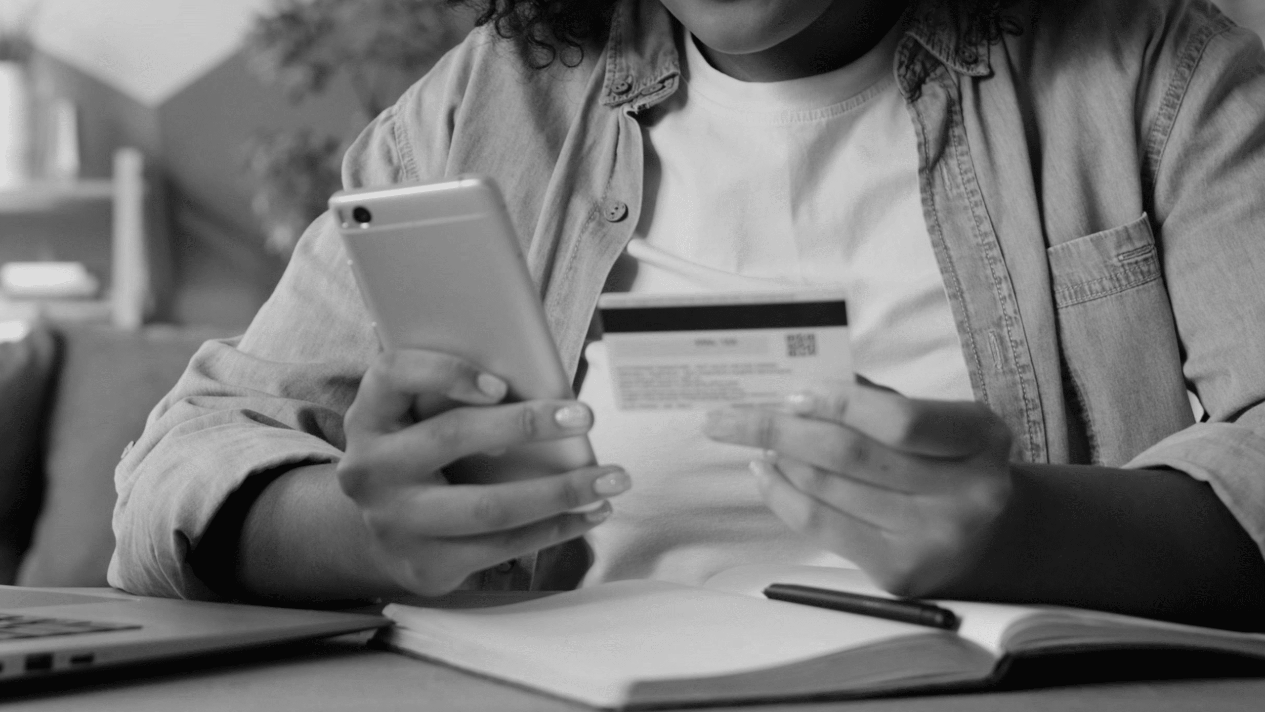 A person holds a smartphone in one hand and a credit card in the other while sitting at a desk with a notebook, pen, and a partially visible laptop.