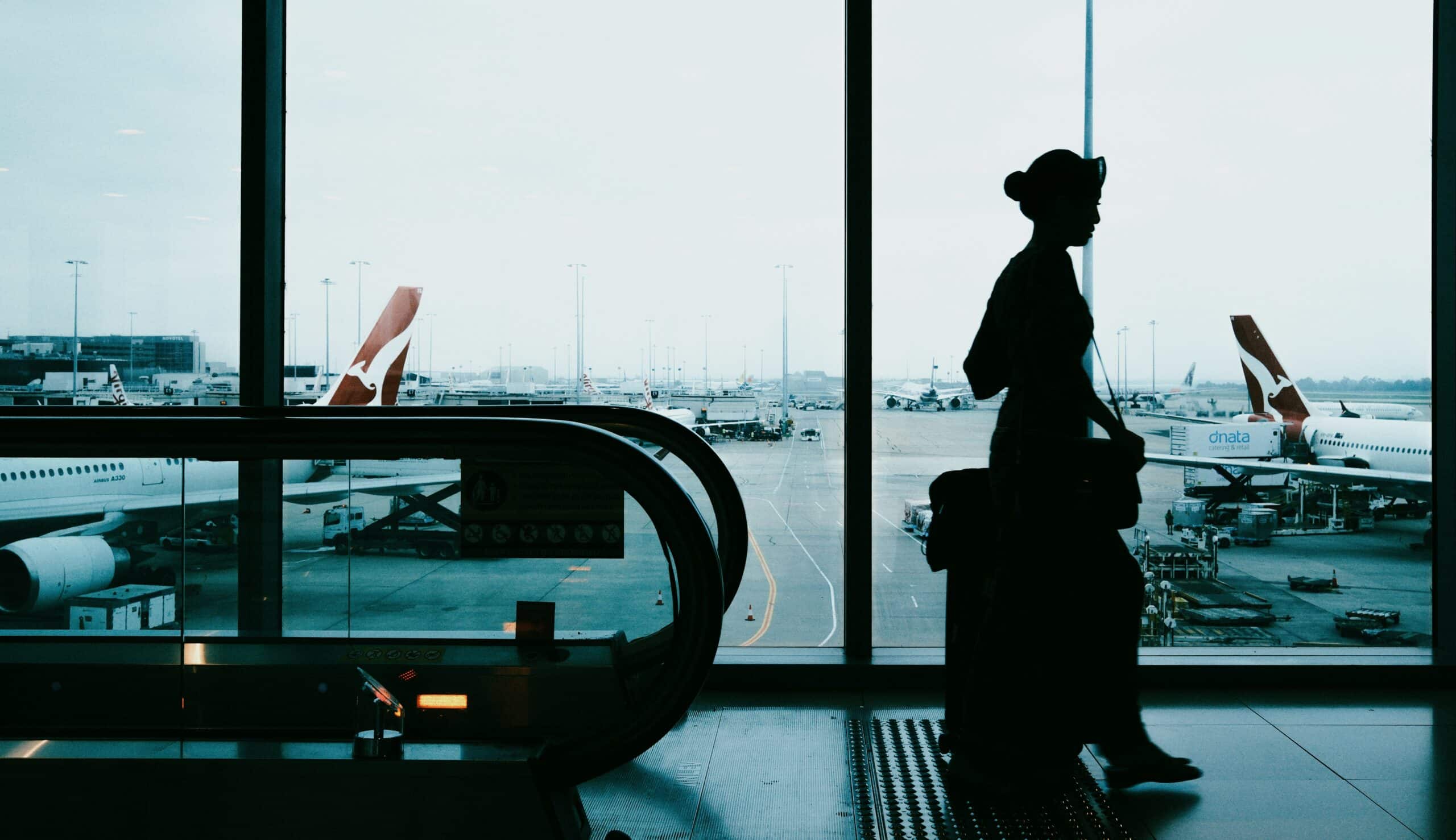 A person with a suitcase stands in silhouette at an airport terminal, near an escalator, with several airplanes visible outside on the tarmac through large windows.