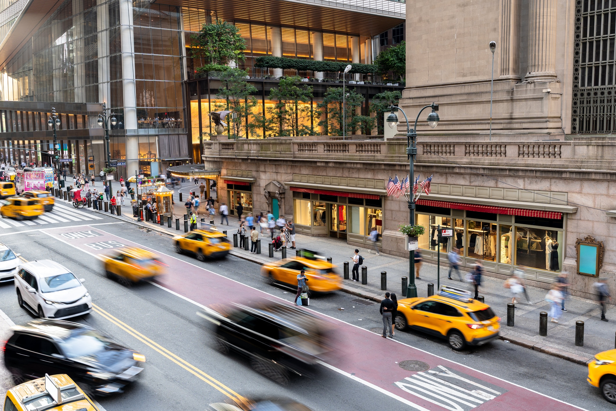 Yellow taxis and other vehicles drive past pedestrians on a busy city street outside a large building with American flags. A modern building with glass and greenery is visible in the background.