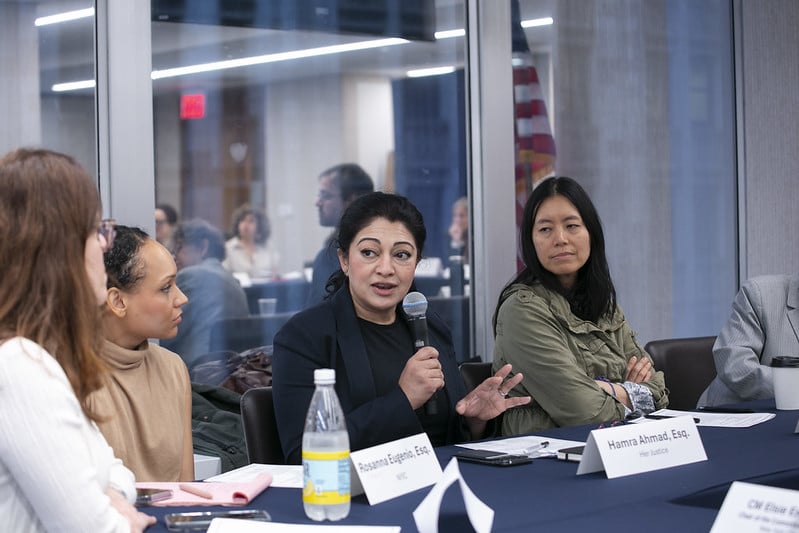 Four women sit at a conference table during a panel discussion. One woman speaks into a microphone while others listen. Papers, a water bottle, and a coffee cup are on the table. People and an American flag are visible in the background.
