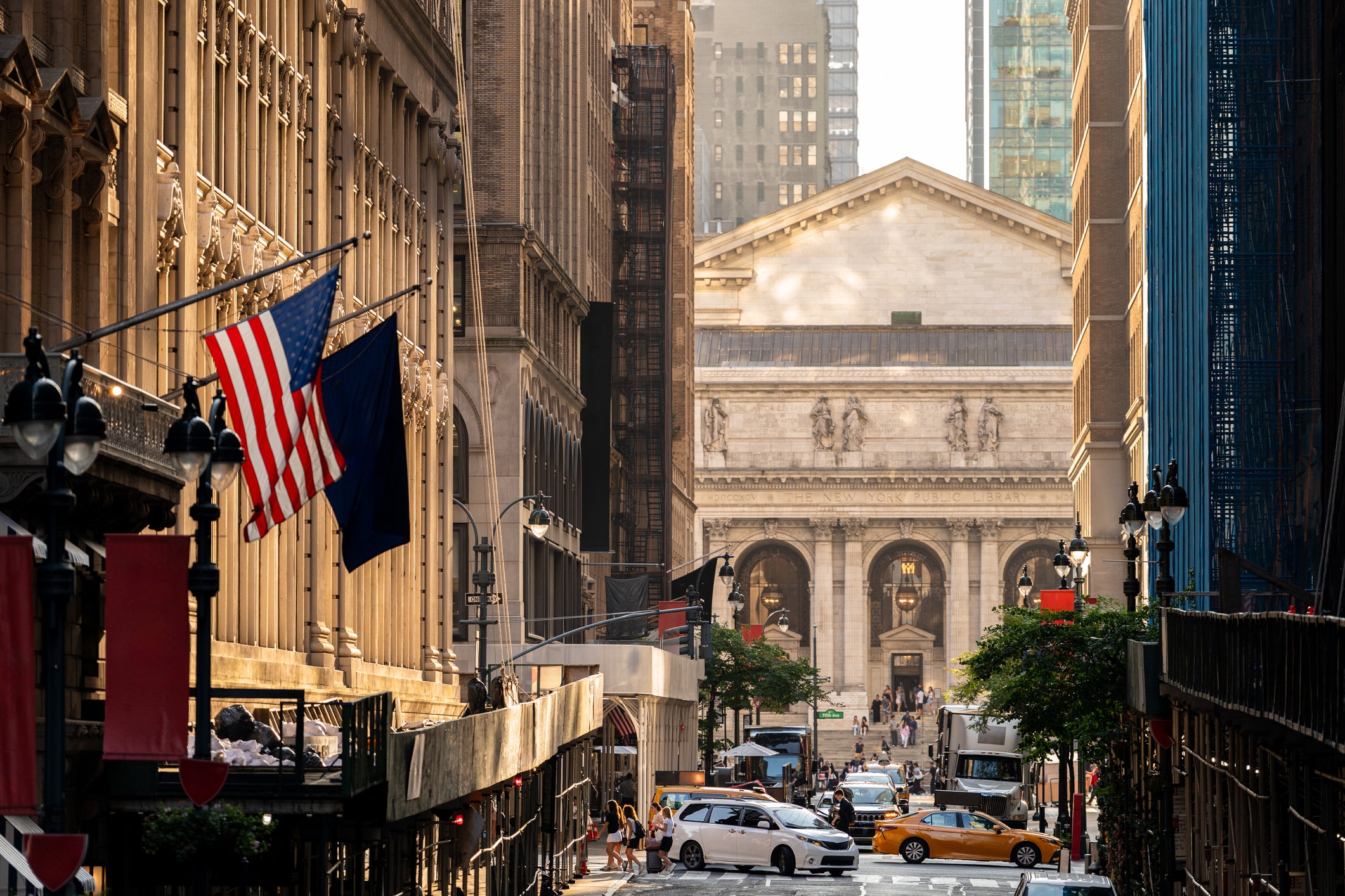 A busy city street with cars, taxis, and people. Two American flags hang from a building on the left. In the background is a large classical building with columns and a peaked roof. Bright daylight illuminates the scene.