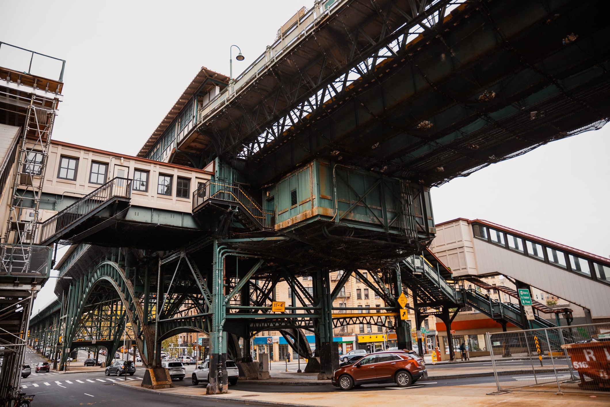 An elevated train station with metal supports spans across an urban street. Cars are parked below, and surrounding buildings and storefronts are visible in the background under an overcast sky.
