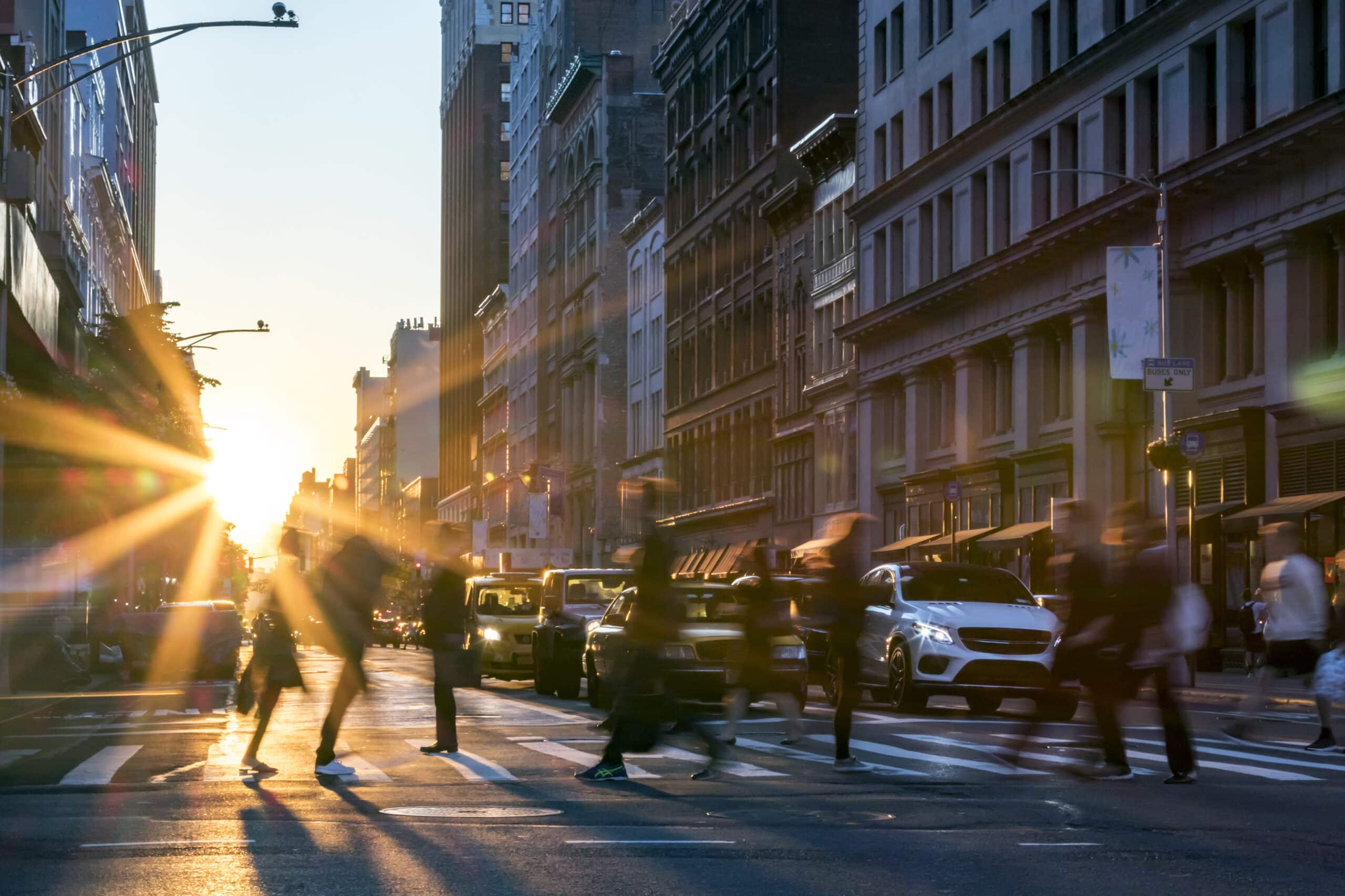 Several people cross a city street at a crosswalk during sunset, with sunlight streaming between tall buildings and cars stopped at the intersection.