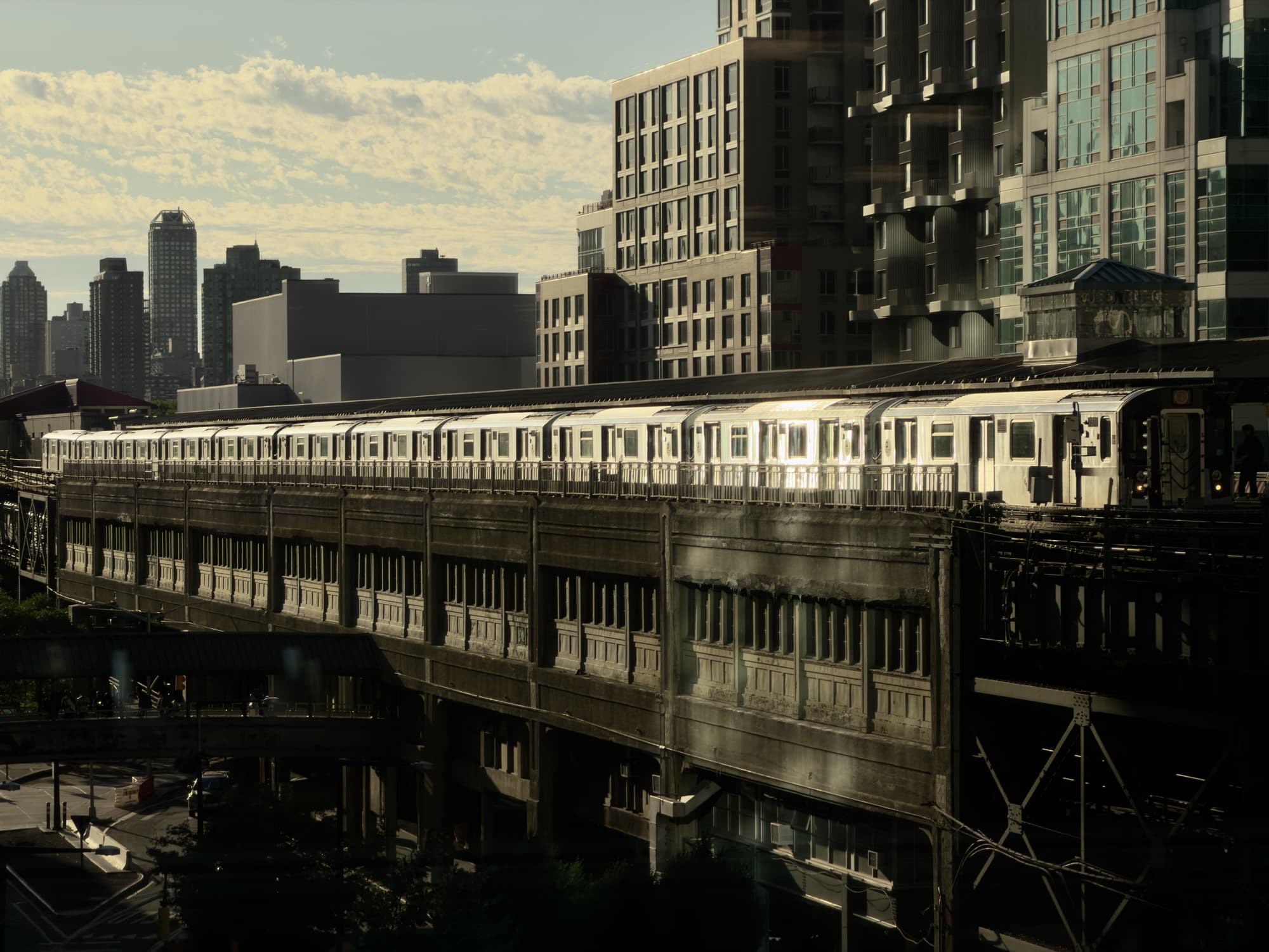 A subway train travels on an elevated track through a city with modern high-rise buildings. The sunlight reflects off the trains surface. The sky is partly cloudy, and the scene has a mix of light and shadow.