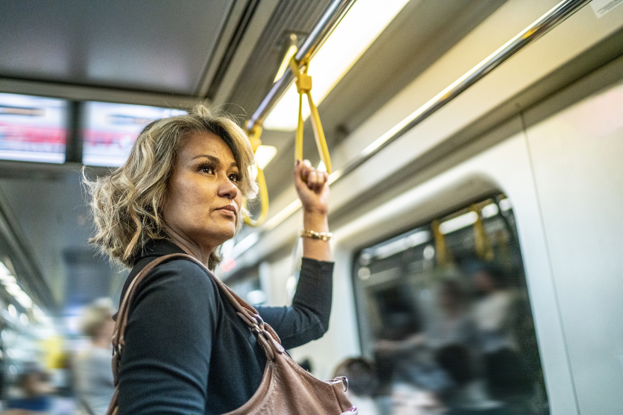 A woman with shoulder-length blonde hair stands on a subway train, holding a yellow strap with her left hand. She wears a brown bag and a black long-sleeve shirt. Other passengers are visible in the background.