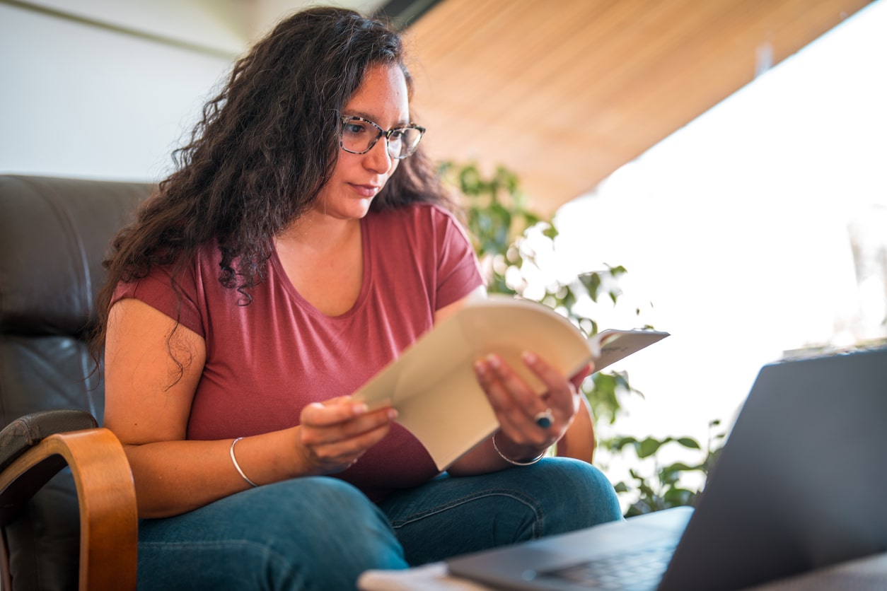 A person with long hair and glasses sits in an armchair, holding and reading a book. There is an open laptop in front of them, and a plant is visible in the background.