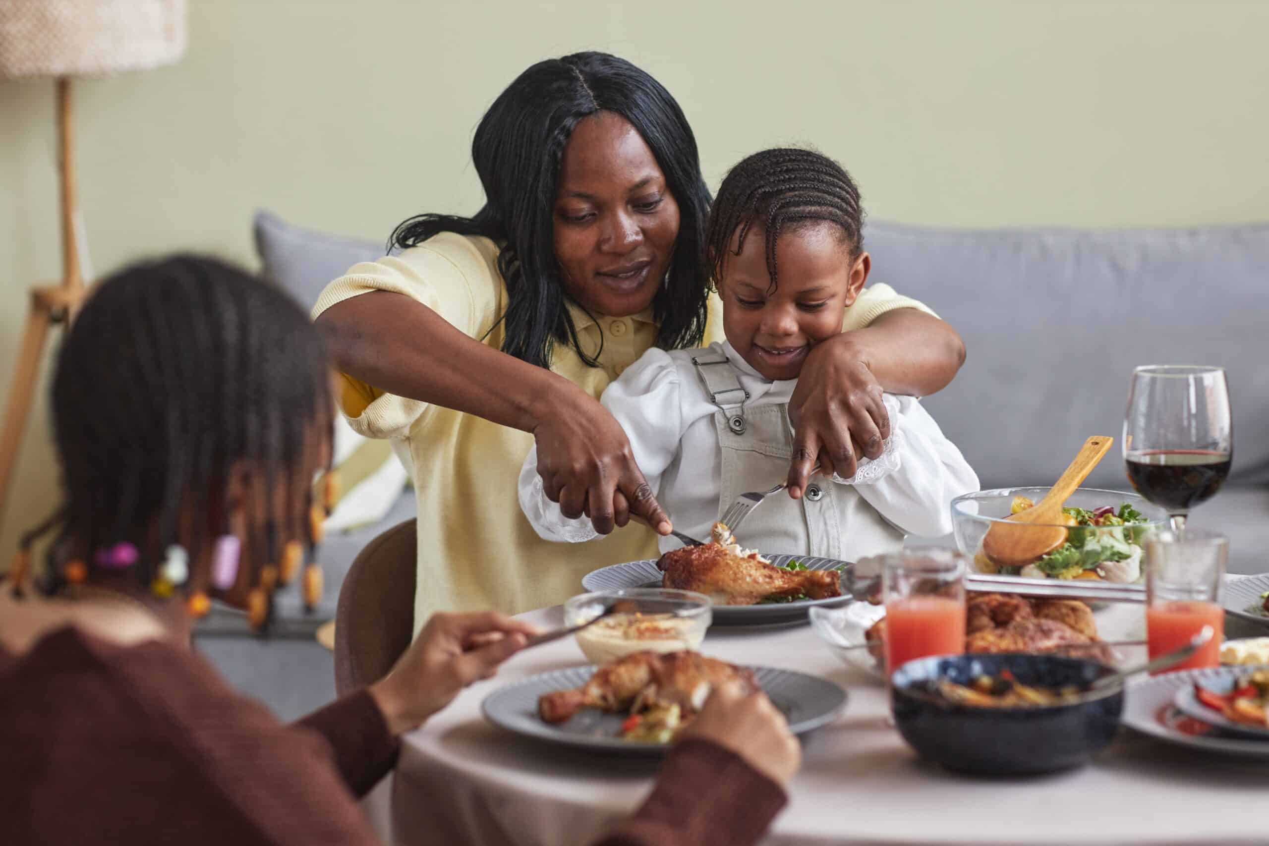 An adult helps a child cut food on a plate during a meal at a table. Another child is seated nearby. The table has plates of food, drinks, and a bowl. The setting appears to be a casual, indoor dining space.