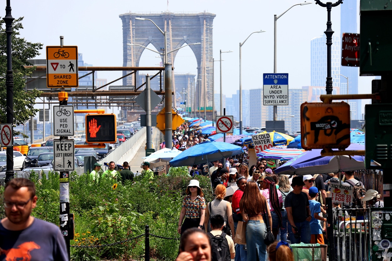 Crowds of people walk near the Brooklyn Bridge on a sunny day. Vendors with umbrellas line the path. Traffic signs, a pedestrian signal, and city buildings are visible in the background.