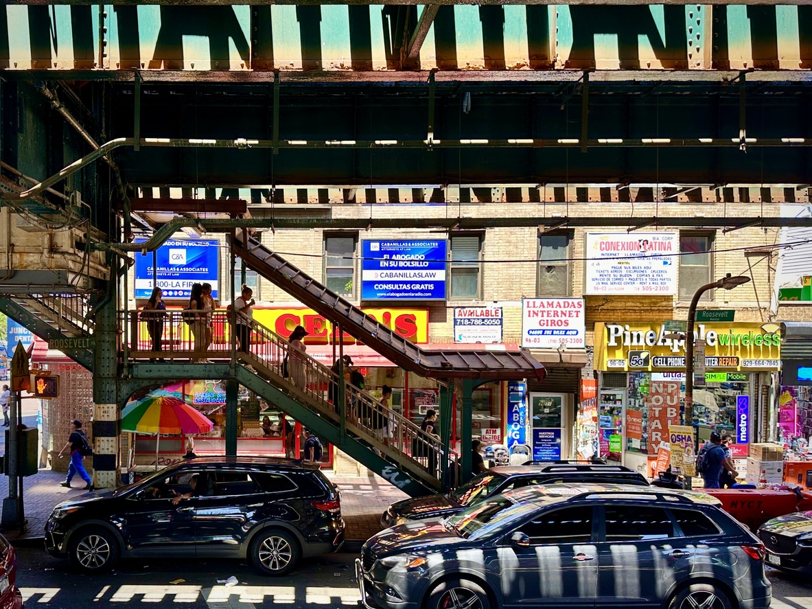 Elevated train tracks and stairway over a busy city street with parked cars below, various colorful storefronts, and people walking on the stairs and sidewalk. Bright sunlight highlights the scene.
