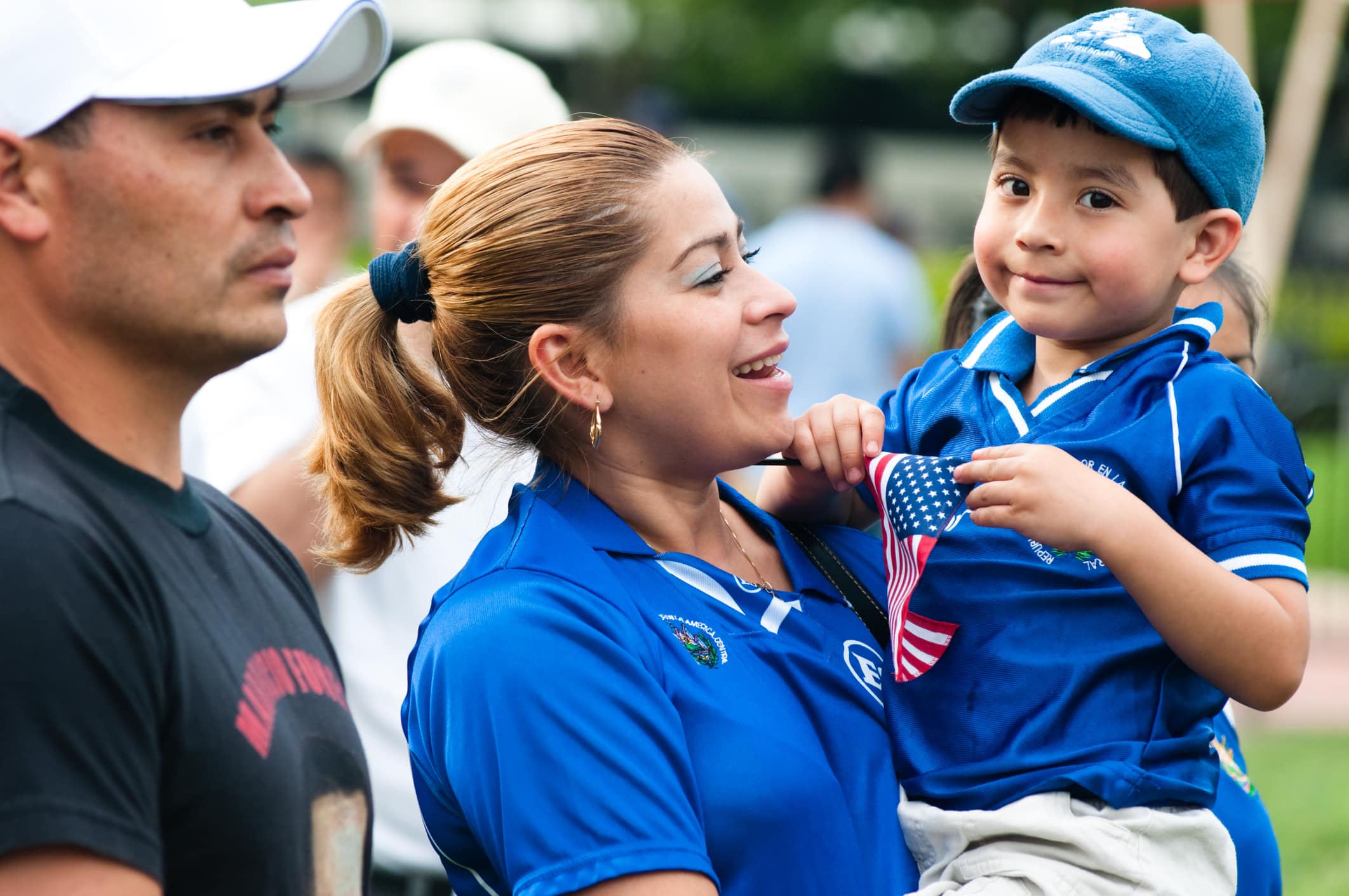 A woman in a blue shirt holds a young boy wearing a blue cap and shirt, who is holding a small American flag. A man in a black shirt and white cap stands nearby. They are outdoors with other people in the background.