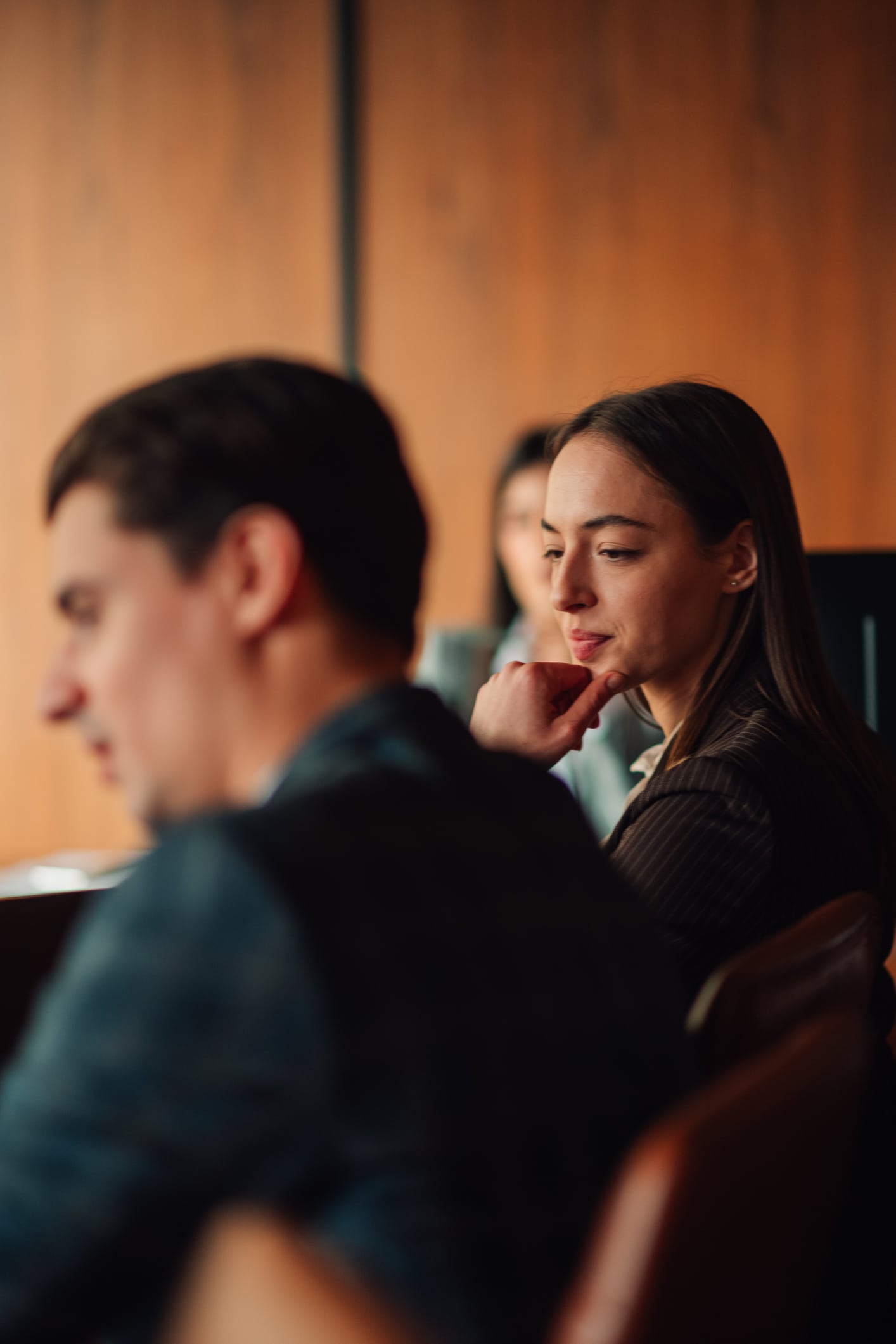 A woman sits at a table in a meeting room, looking thoughtful with her hand near her chin. Two other people are partially visible, one in the foreground and one in the background. The setting appears professional.