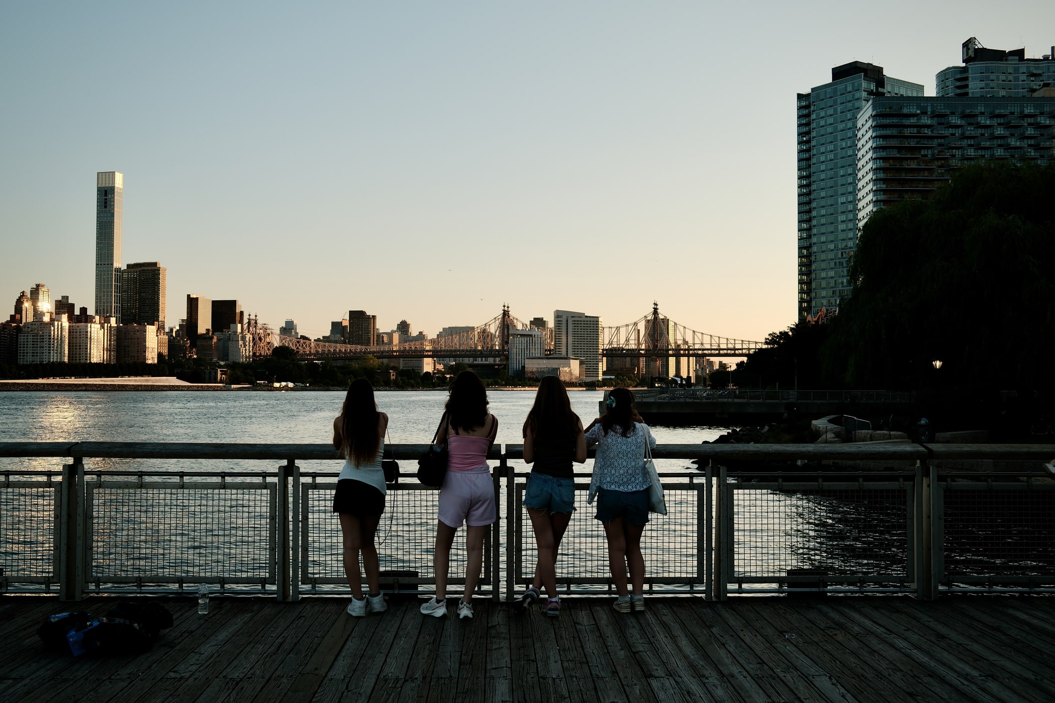 Four people stand on a wooden boardwalk overlooking a river at sunset, with city buildings and a bridge visible in the background.
