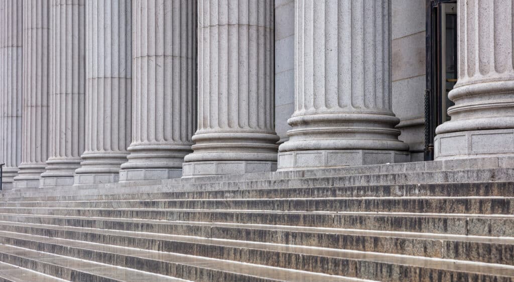 A row of large stone columns stands behind a series of wide, shallow steps outside a classical-style building. The columns are evenly spaced and the image shows their bases and shafts.