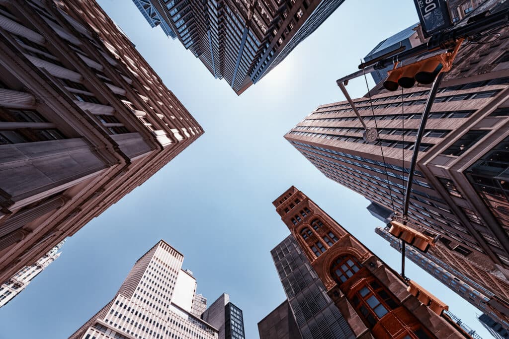 Skyscrapers and tall buildings surround the view, shot from below looking up toward a clear blue sky. A traffic light and street sign are visible in the lower right corner.