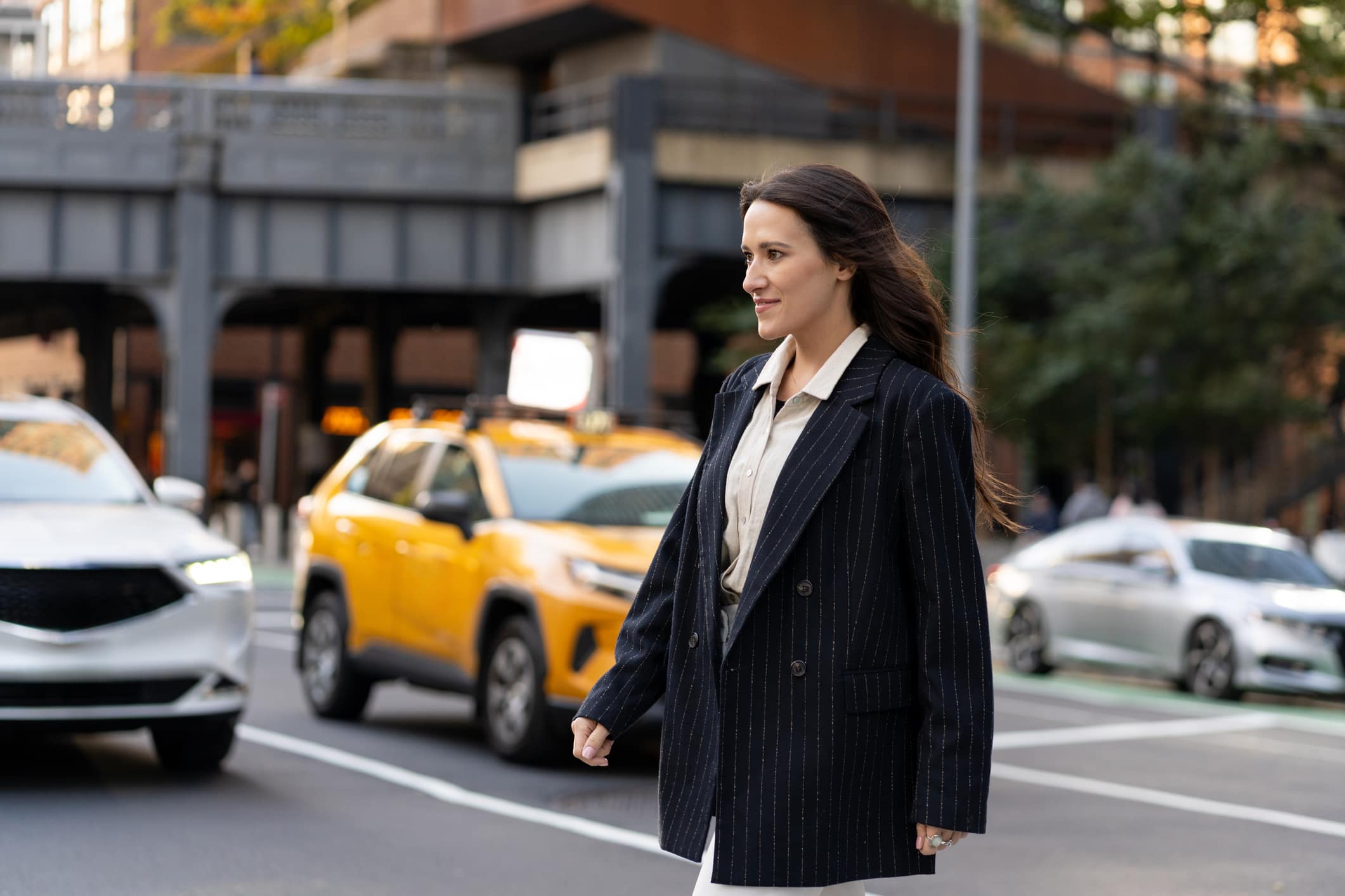 A woman wearing a dark pinstripe blazer walks across a city street. Yellow and white cars wait behind her. Urban buildings and greenery are visible in the background.