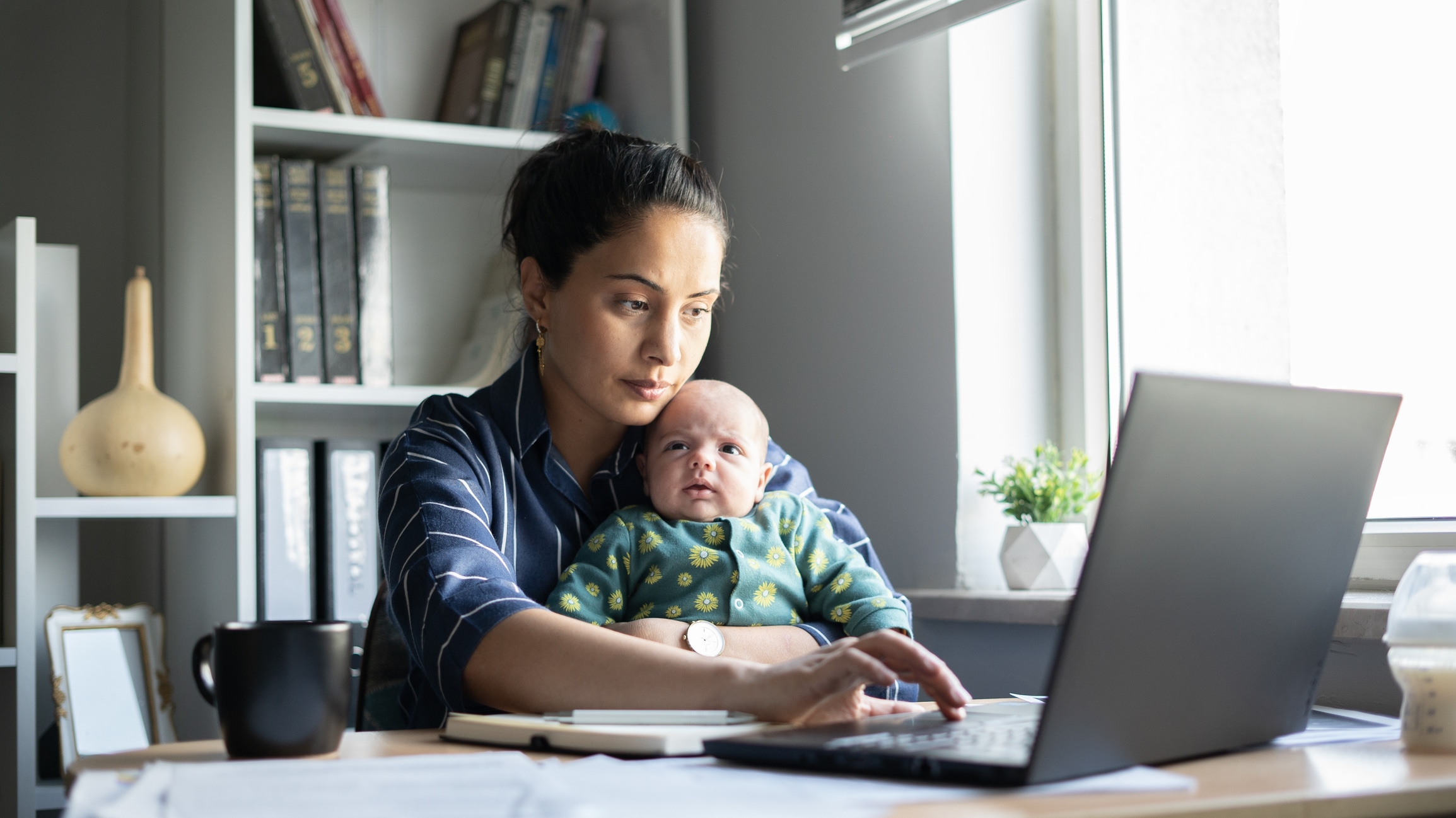 A woman sits at a desk working on a laptop while holding a baby. Papers, a mug, and a bottle are on the desk. Shelves with books and a plant are in the background. The scene is lit by natural light from a window.