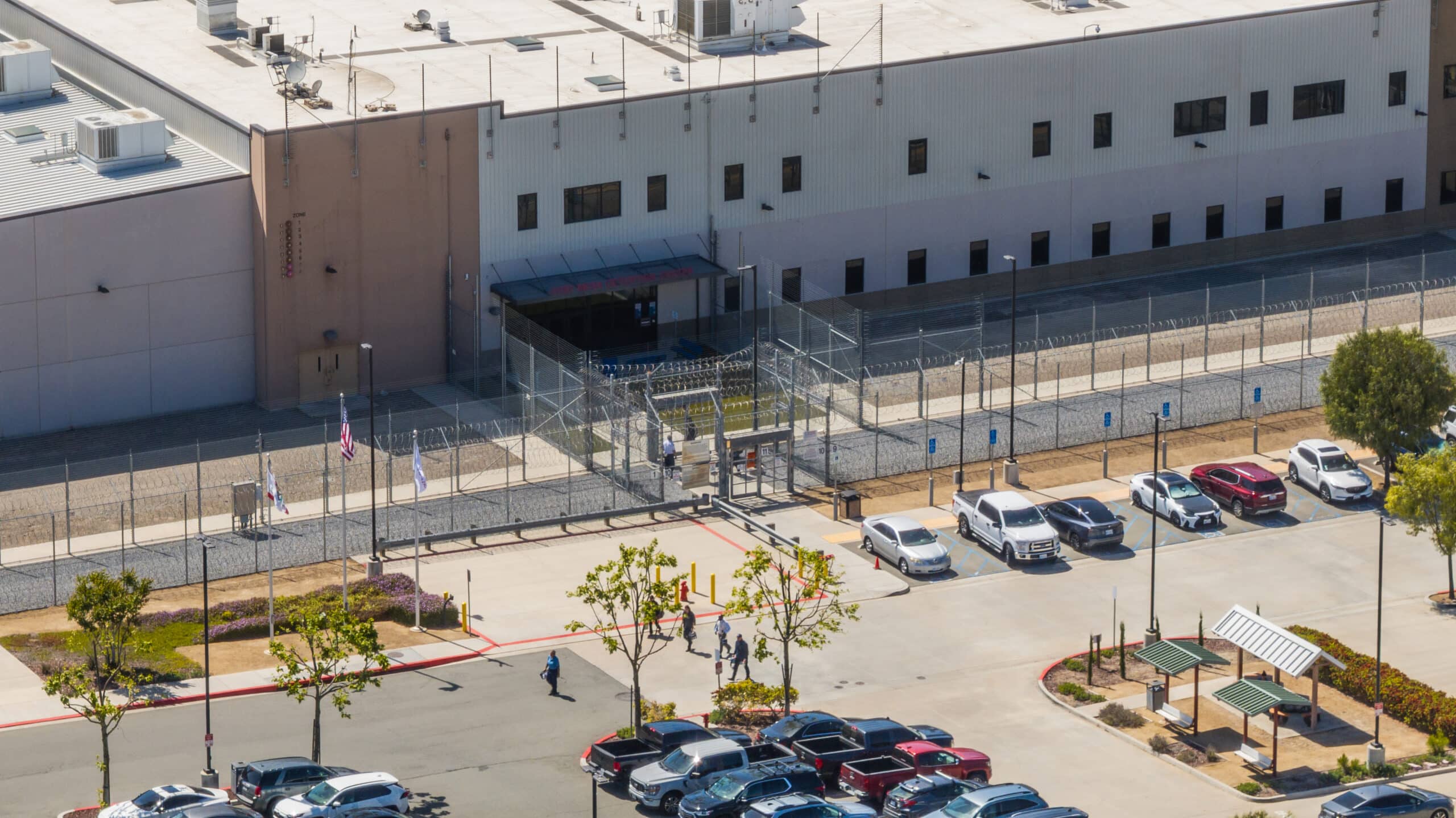 Aerial view of a fenced facility with armed security, several parked cars, people walking nearby, and a parking lot with trees and covered benches in the foreground.