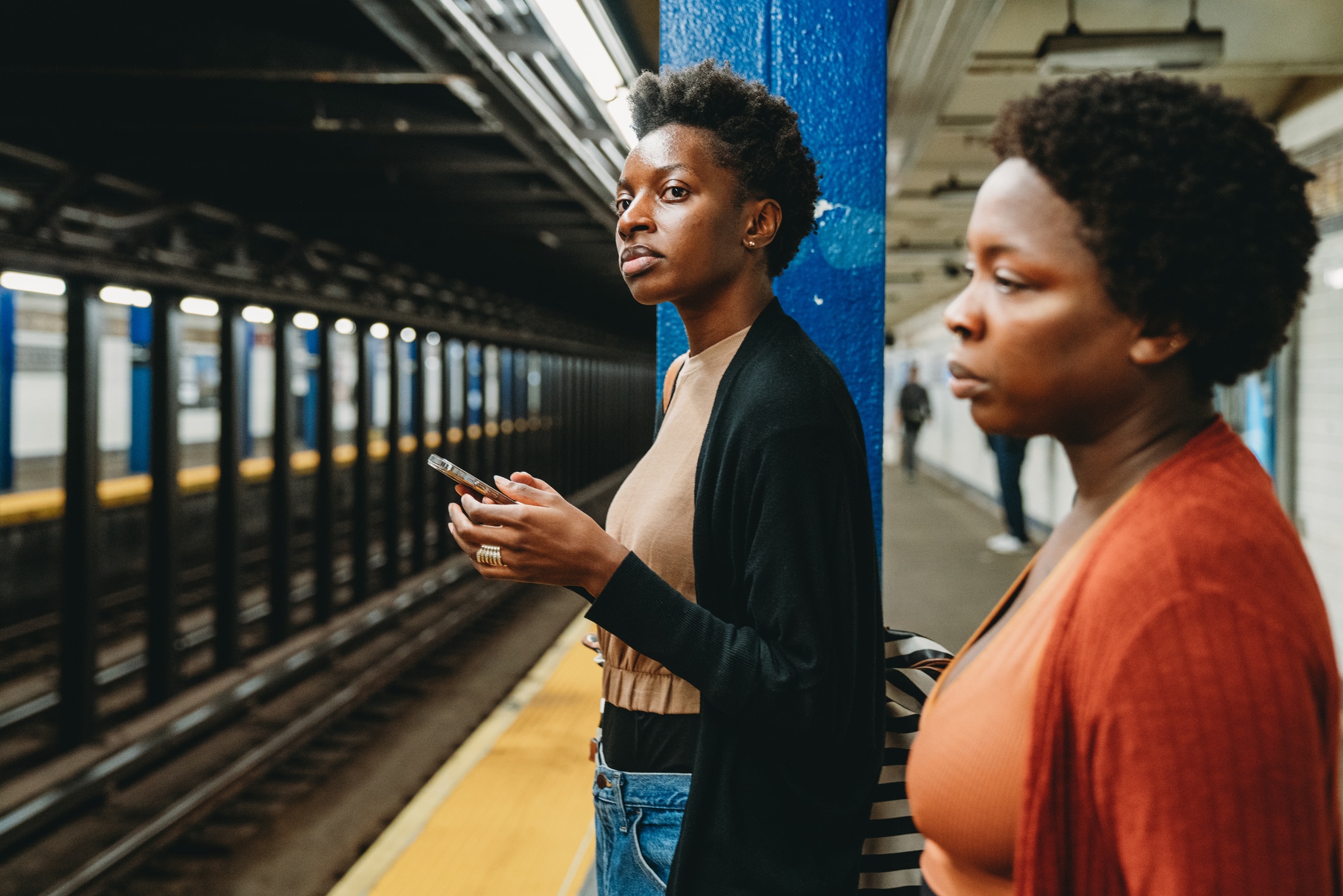 Two women stand on a subway platform next to a blue column. One holds a phone and looks ahead, while the other gazes toward the tracks. The subway rails and tunnel are visible in the background.